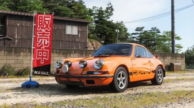 An older Japanese coupe sitting outside a rural used car lot deep in the countryside.